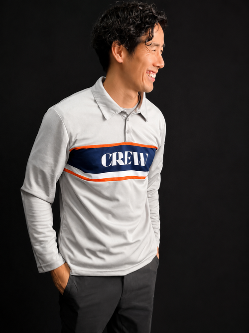 A man wearing a light gray long-sleeve polo shirt with a navy blue chest stripe outlined in orange and the word 'CREW' in white, paired with dark gray pants, set against a solid black background.