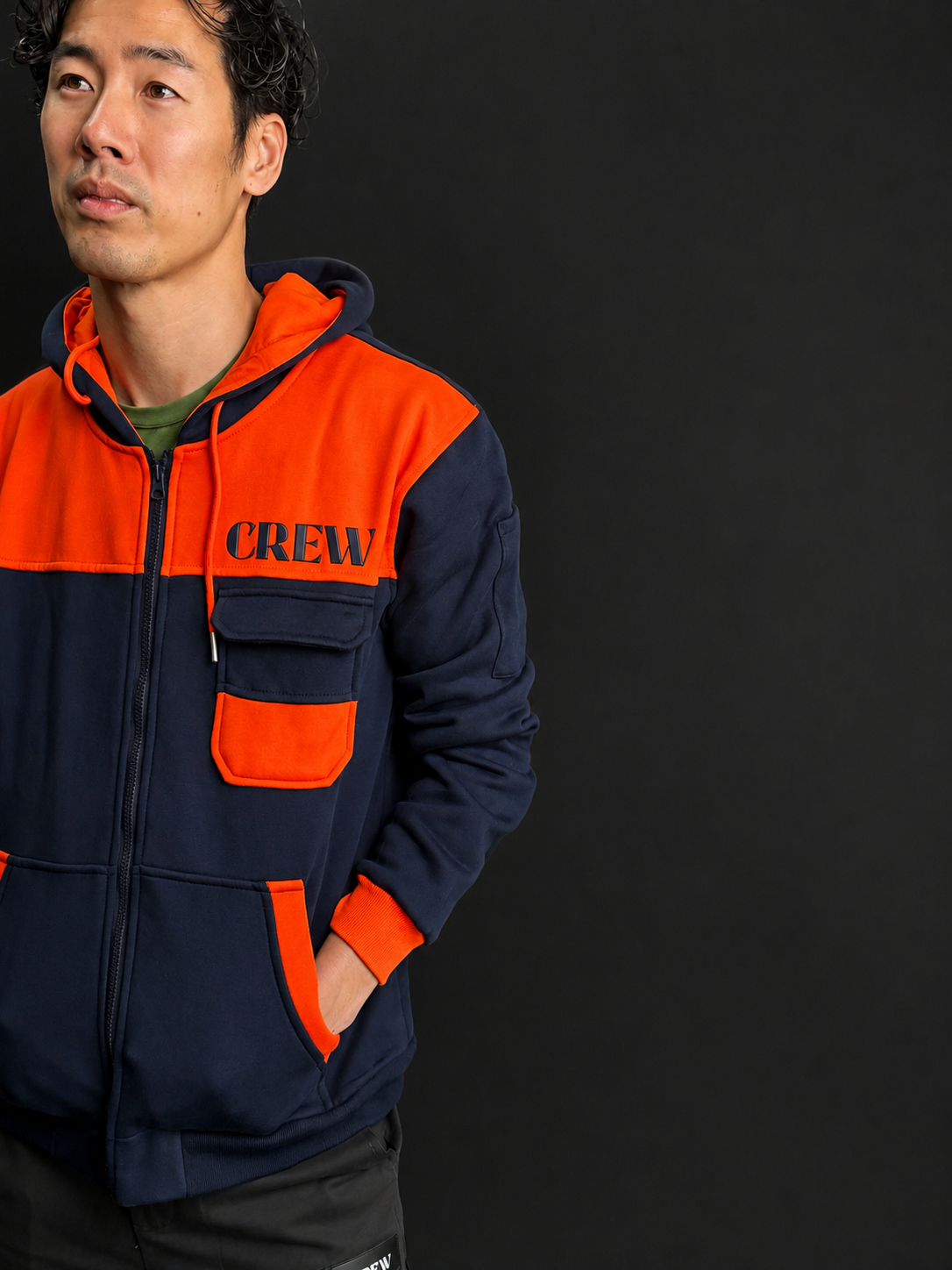 A man wearing a color-blocked zip-up hoodie with a navy blue body, bright orange hood and chest pocket, and 'CREW' printed in black on the chest, set against a dark studio background.