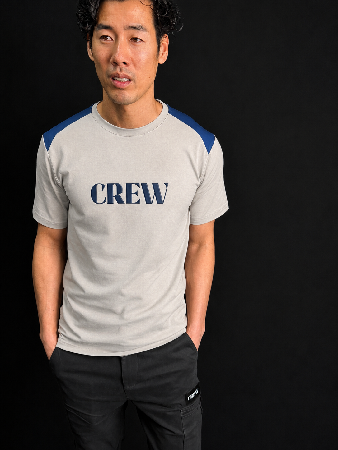 A man wearing a light gray crew-neck t-shirt with blue shoulder panels and a navy blue 'CREW' logo on the chest, paired with dark gray pants, set against a solid black background.