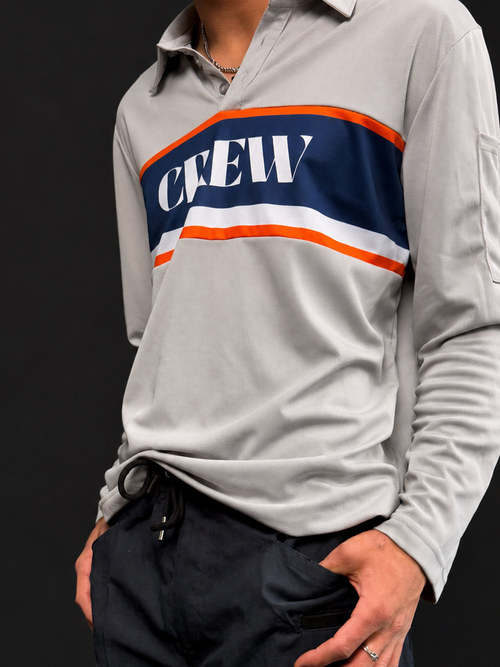 A man wearing a light gray long-sleeve crewneck shirt with a navy blue chest stripe featuring orange and white details, paired with dark navy shorts that have a white 'CREW' logo patch on the lower left leg.